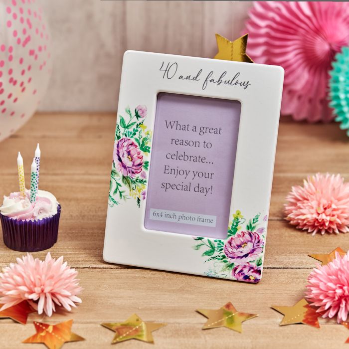 40 and fabulous photo frame with floral decoration, displayed on a wooden surface with birthday decorations including a cupcake, candles, paper flowers, and gold star confetti.