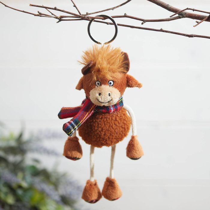 Plush Highland cow keyring with a tartan scarf, hanging from a tree branch with a blurred natural background. 