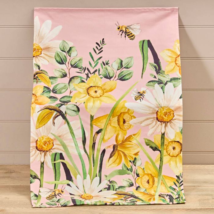 Floral tea towel with yellow daffodils, white daisies, green foliage, and bees on a pink background, displayed against a wooden panel wall. 
