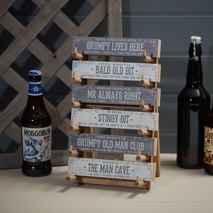 Humorous wooden signs with phrases like 'Grumpy Lives Here' and 'The Man Cave' displayed on a stand, surrounded by beer bottles, in a rustic setting.
