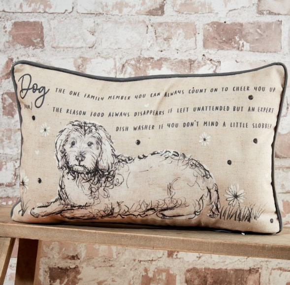 Rectangular cushion with a sketch-style illustration of a dog and playful text about dogs, displayed on a wooden bench against a rustic brick wall.