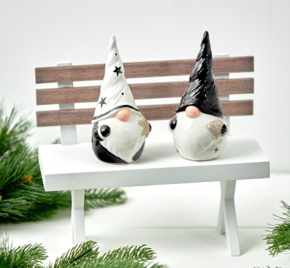 Two black and white gonks sitting on a white bench with wooden backing, surrounded by fresh green foliage and evergreen branches.