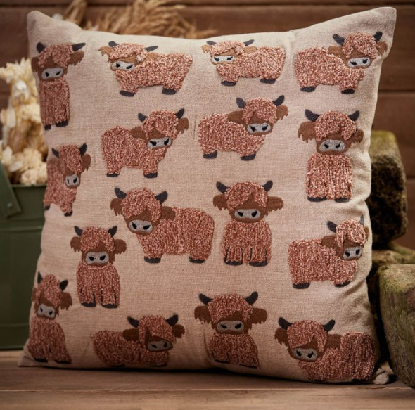 Beige cushion decorated with a repeated pattern of cute, textured highland cow motifs in warm brown tones, styled in a rustic wooden interior setting.