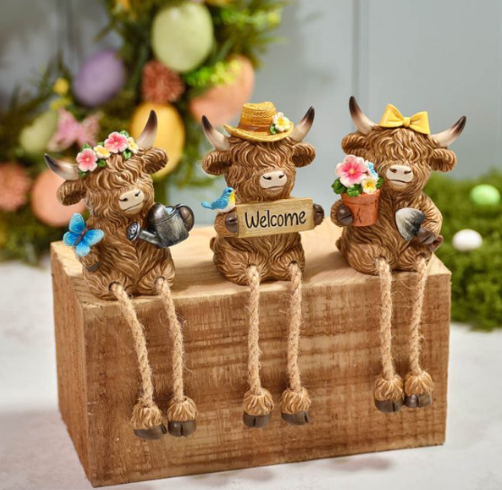 Three textured Highland cow figurines with flowers and accessories sitting on a wooden block, with colourful decorative eggs in the blurred background. 