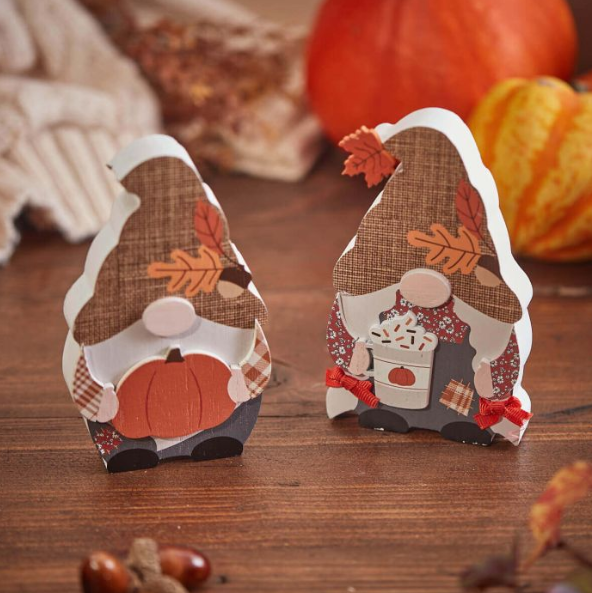 Two autumn-themed gonk ornaments standing on a wooden surface, one holding a pumpkin and the other a mug, surrounded by pumpkins and cosy fall décor.